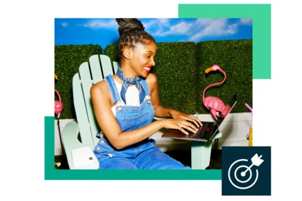 ChatGPT said: Smiling woman in denim overalls types on a laptop while sitting outside on a patio, surrounded by pink flamingos and green hedge walls.